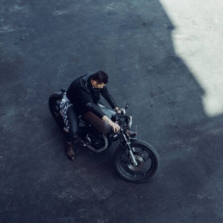 Top view of a handsome rider guy in black biker jacket, checkered shirt, denim and boots ride his classic style cafe racer motorcycle. Bike custom made in vintage garage. Brutal fun urban lifestyle.の写真素材
