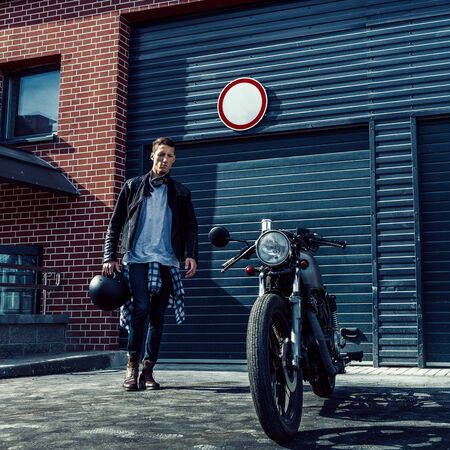 Handsome young rider guy in black biker jacket and jeans go to his classic style cafe racer motorcycle industrial gates as background. Bike custom made in vintage garage. Brutal fun urban lifestyle.の写真素材