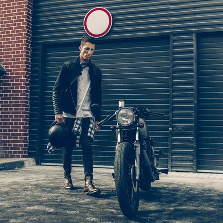 Handsome young rider man in black biker jacket and boots go to his classic style cafe racer motorcycle industrial gates as background. Bike custom made in vintage garage. Brutal fun urban lifestyle.の写真素材