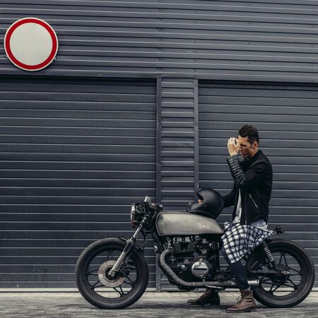 Handsome rider man biker correct hair after helmet sit on classic style cafe racer motorcycle near industrial gates. Bike custom made in vintage garage. Brutal fun urban lifestyle. Outdoor portrait.の写真素材