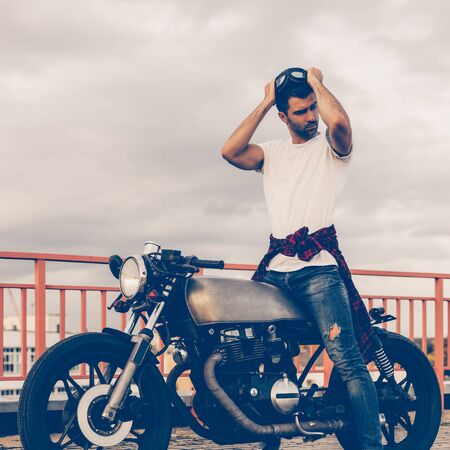 Handsome rider man with beard and mustache posing while take off protection glasses near classic style biker cafe racer motorcycle. Bike custom made in vintage garage. Brutal fun urban lifestyle.の写真素材