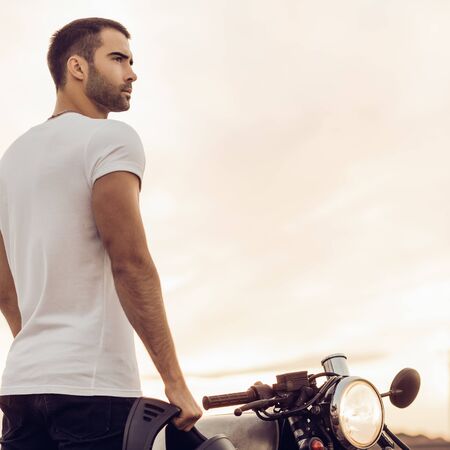 Sporty biker handsome rider man in white t-shirt go travel on classic style cafe racer motorbike on rooftop at sunset. Vintage bike custom made in garage. Brutal urban lifestyle. Outdoor portrait.の写真素材