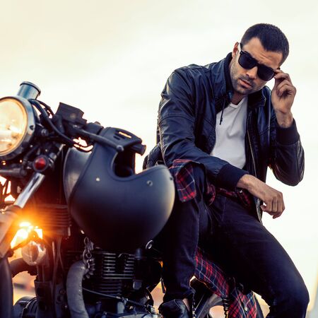Handsome rider guy with beard and mustache in black biker jacket take off sunglasses on classic style cafe racer motorcycle at sunset. Bike custom made in vintage garage. Brutal fun urban lifestyle.の写真素材