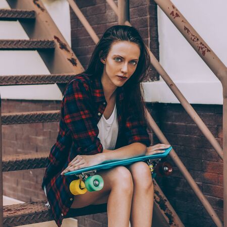 Young beautiful girl in checkered hipster shirt, short jeans shorts and white sneakers sit on the stairs with blue mini skate board longboard. Urban scene, city life. Cute sexy lady look to camera.の写真素材
