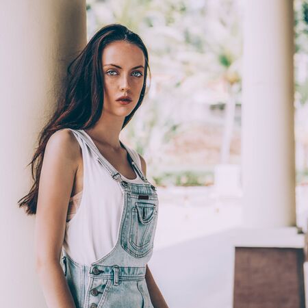 Portrait of slim beautiful girl in white t-shirt and short jeans overall. Urban scene, city life. Cute attractive sexy hipster lady standing in front of a column, posing and looking to the camera.の写真素材