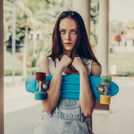 Close-up portrait of sexy beautiful woman in white t-shirt and short denim overall holding short skate board. Urban scene, city life. Cute attractive sexy hipster lady standing in front of a column.の写真素材