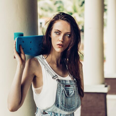 Portrait of sexy beautiful woman in white t-shirt and short jeans overall holding blue short skate board. Urban scene, city life. Cute attractive sexy hipster lady standing in front of a column.の写真素材