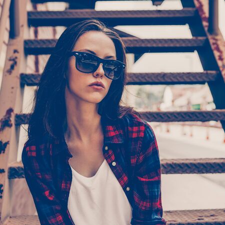 Closeup portrait of young beautiful girl in checkered hipster shirt, short jeans shorts, white sneakers and sunglasses sit on the stairs. Urban scene, city life. Cute sexy woman look to the camera.の写真素材