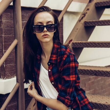 Close-up portrait of young beautiful girl in checkered hipster shirt, white t-shirt and trendy sunglasses sit on the stairs. Urban scene, city life. Cute attractive sexy woman looking to the camera.の写真素材