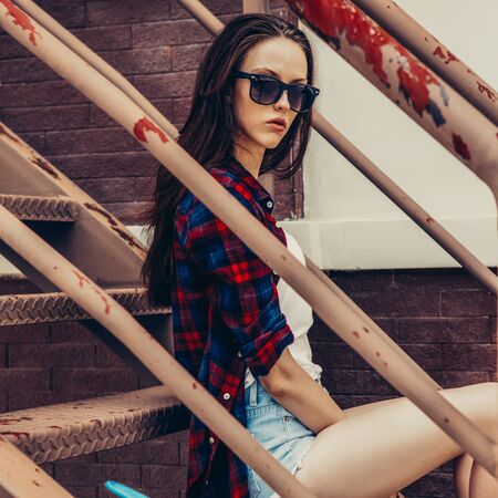 Young beautiful girl in checkered hipster shirt, short jeans shorts, white sneakers and sunglasses sit on the stairs with mini skate board. Urban scene, city life. Cute sexy lady look to the camera.の写真素材