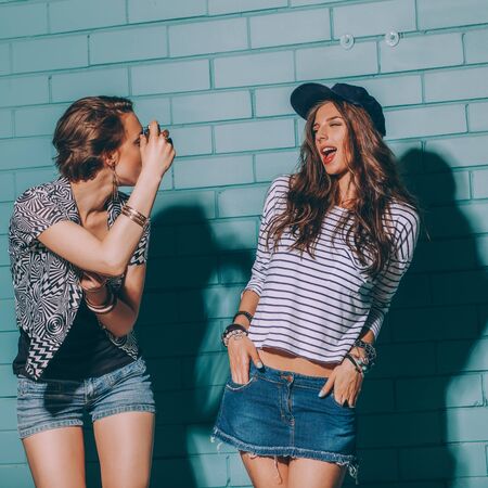 Woman in stylish bright outfit and denim shorts going crazy and having great time. Standing together near blue brick wall with photo camera and have fun while taking selfie portrait of each other.の写真素材