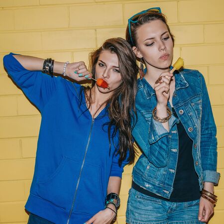 Lifestyle portrait of two beautiful best friends hipster woman wearing stylish bright outfits and having great time. Standing near yellow brick wall enjoying day off, have fun and eating ice-cream.の写真素材