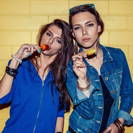 Lifestyle portrait of two beautiful best friends hipster girls wearing stylish bright outfits and having great time. Standing together near yellow brick wall enjoying ice-cream and look to the camera.の写真素材