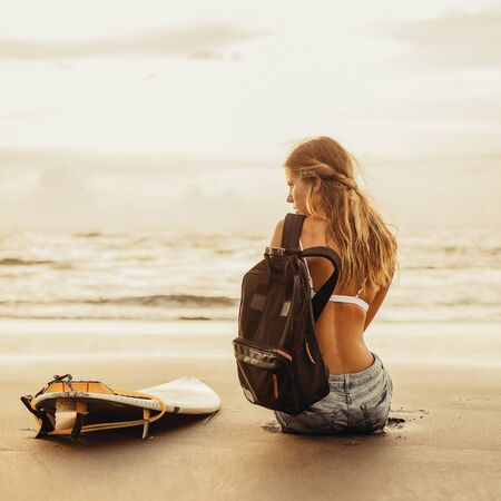 Young surfer girl in sexy bikini and jeans shorts with surf board and travel backpack at sunset ocean. Modern family lifestyle, people water sport adventure camp and extreme swim on summer vacationの写真素材