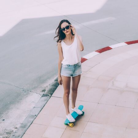 Slim sporty young beautiful woman in white t-shirt, short jeans shorts and trendy sunglasses ride blue shortboard penny skateboard. Urban scene, city life. Cute attractive sexy hipster lady have fun.の写真素材