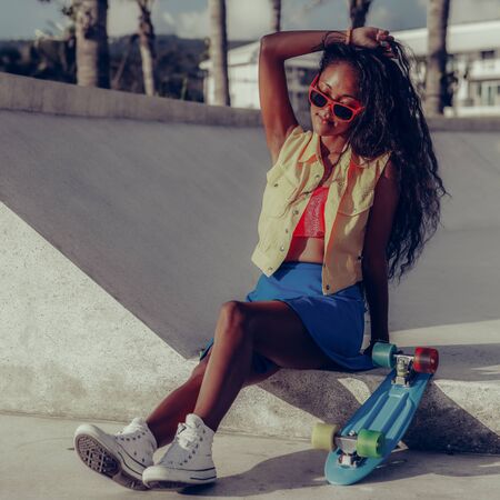 Outdoor lifestyle portrait of black young female in bright outfit. Young hipster woman sitting with skateboard and touch her long beautiful curly hair. She is happy. Sunny summer day. Swag, fashion.の写真素材