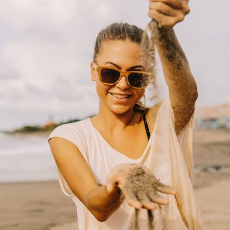 Pretty girl in casual clothes and sunglasses watch the sand running through hand and smile. Lady on sea beach sunset or ocean sunrise. Travel, explore, active yoga and meditation lifestyle concept.の写真素材