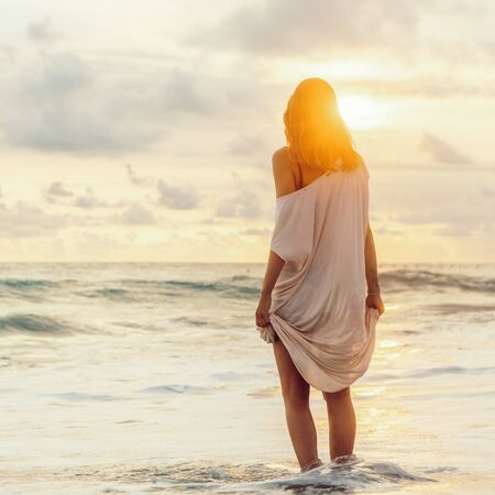 Hipster girl after hot summer day hold her long light dress trying not to wet it in cool water. Fit lady in sand sea beach sunset or ocean sunrise. Travel, active, yoga, freedom lifestyle concept.の写真素材