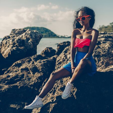 Outdoor lifestyle portrait of a black sexy  in blue skirt, white sneakers, pink bra and sunglasses. Hipster  sitting at big rock at stone beach. Sunny hot summer day. Swag, fashion.の写真素材