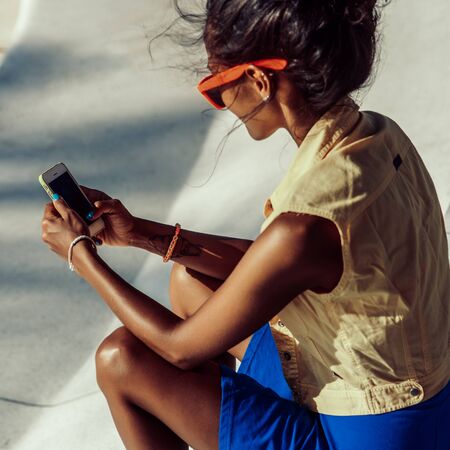 Outdoor lifestyle portrait of black young female in bright outfit and sunglasses. Hipster girl sitting at skate park with blue penny longboard skateboard and texting on the smartphone. Swag, fashion.の写真素材
