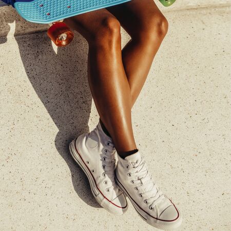 Outdoor lifestyle portrait of black young lady in bright outfit. Hipster girl sitting at skate park with blue penny shortboard skateboard and chatting on the smartphone phone. Swag, fashion. Sunny dayの写真素材