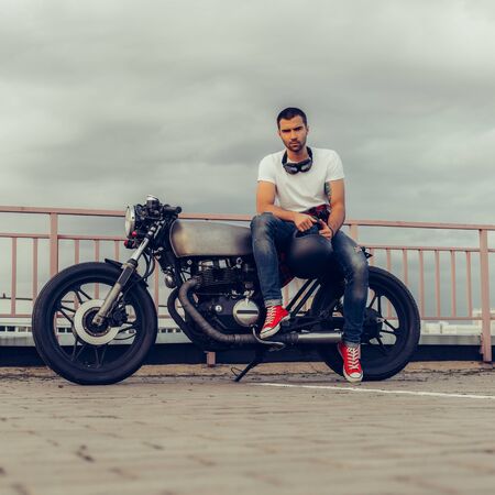 Rider guy with beard and mustache in red sneakers and black googles sit on classic style biker cafe racer motorcycle. Bike custom made in vintage garage. Brutal fun urban lifestyle. Outdoor portrait.の写真素材