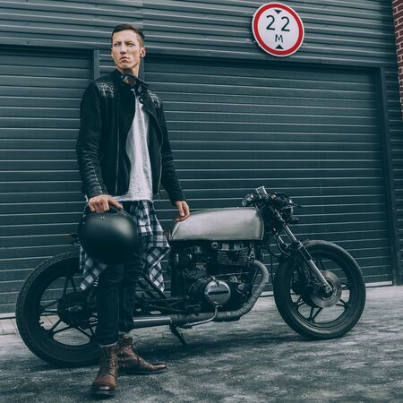 Handsome rider man in black leather biker jacket and boots near classic style cafe racer motorcycle industrial gates as background. Bike custom made in vintage garage. Brutal fun urban lifestyle.の写真素材