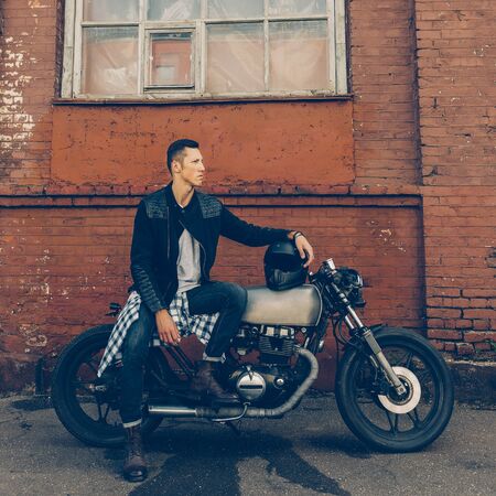 Handsome biker guy in black leather jacket sit on classic style cafe racer motorcycle and hold helmet on gas tank. Bike custom made in vintage garage. Brutal fun urban lifestyle. Outdoor portrait.の写真素材