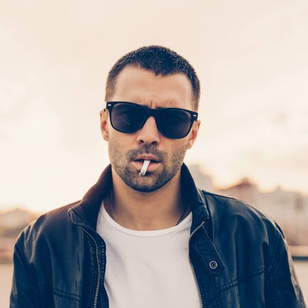Close up of a handsome rider man with beard and mustache in black biker jacket, fashion sunglasses smoking cigaret near classic style cafe racer motorbike at sunset. Brutal urban lifestyle portrait.の写真素材