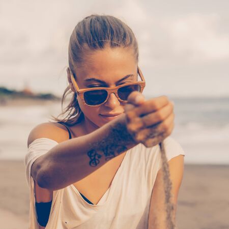 Pretty woman in casual clothes and wooden sunglasses look to the sand running through hand. Lady on sea beach sunset or ocean sunrise. Travel, explore, active yoga and meditation lifestyle concept.の写真素材