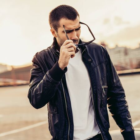 Handsome rider guy with beard and mustache in black biker jacket, white t-shirt and sunglasses smoking cigaret near classic style cafe racer motorbike on rooftop at sunset. Brutal fun urban lifestyle.の写真素材