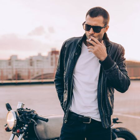 Beautiful happy rider man with beard and mustache in black biker jacket, jeans and fashion sunglasses smoking cigaret near classic style cafe racer motorbike at sunset. Brutal fun urban lifestyle.の写真素材