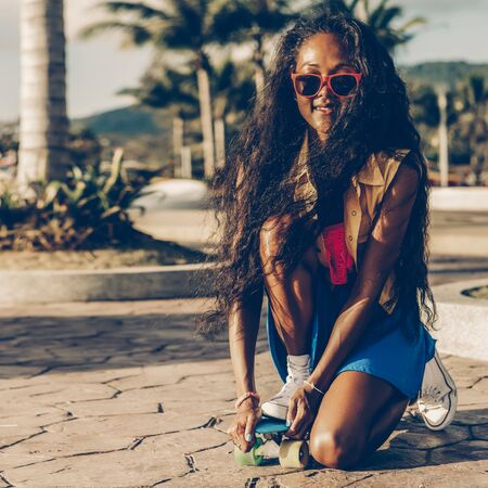 Outdoor lifestyle portrait of sporty black female in bright outfit. Young hipster woman try to ride her blue penny skateboard. She is happy and smile to the camera. Sunny summer day. Swag, fashion.の写真素材