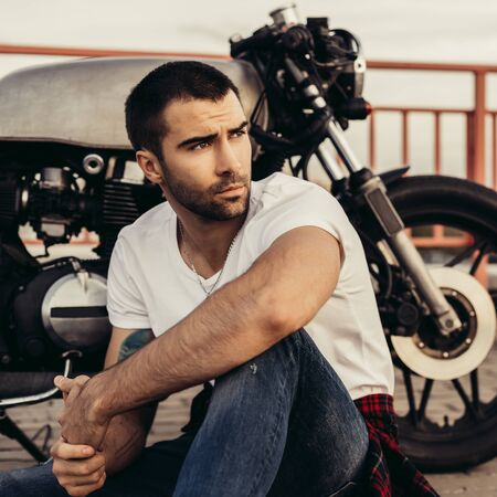 Rider man with beard and mustache in white t-shirt and denim sit near his classic style biker cafe...