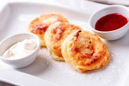 cheesy pancakes sprinkled with powdered sugar close-up on white dish with bowls of sour cream and...