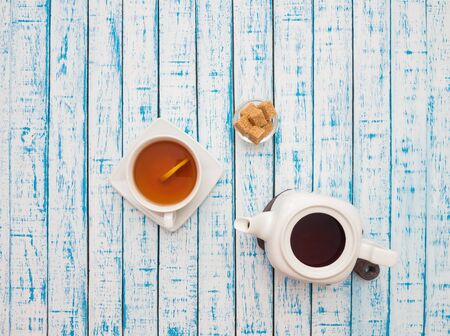 White cup of black tea with lemon, white kettle and cane sugar on a wooden tableの写真素材