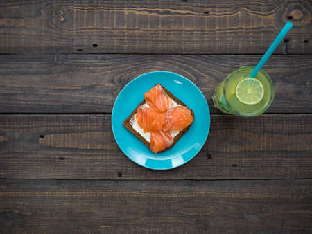 Sandwich with salmon, avakado, cream cheese, black bread in a plate and a large glass of lemonade on a wooden tableの写真素材