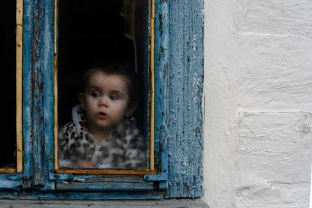 A little girl sits in an old hut on a window on which the paint faded 2018の写真素材