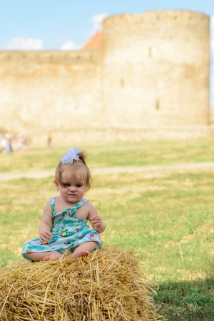 A little girl sits on a straw in a dress and a bow near the wallの写真素材