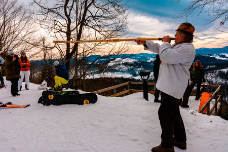 Bukovel ski resort 2021. Grandfather in an embroidered shirt plays the trembita for tourists against the backdrop of the snow-capped Carpathian mountains. Ivano-Frankivsk, Ukraine - February 3, 2019.のeditorial素材