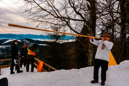 Bukovel ski resort 2021. Grandfather in an embroidered shirt plays the trembita for tourists against the backdrop of the snow-capped Carpathian mountains. Ivano-Frankivsk, Ukraine - February 3, 2019.のeditorial素材