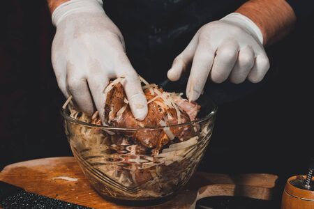 Chef in gloves kneads meat on a plate for cooking kebabs in Georgian. Pork marinade with grated potatoes and onions on a dark background 2021.の写真素材