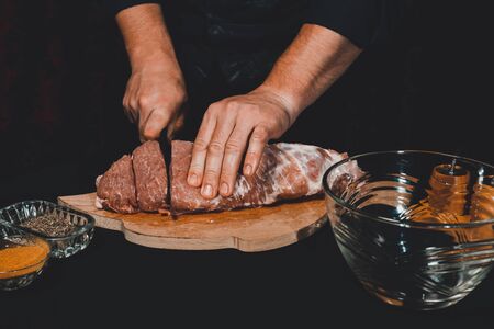 The chef of the restaurant in a dark uniform cuts the meat with a black kitchen knife. Preparation of pork marinated steak to order 2021.の写真素材