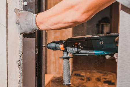 The carpenter drills holes in the wall and the metal door frame with a drill, later screws the...