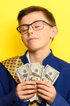 A boy in glasses and an oversized suit with a satisfied face is holding US dollars, a child and big money.の写真素材