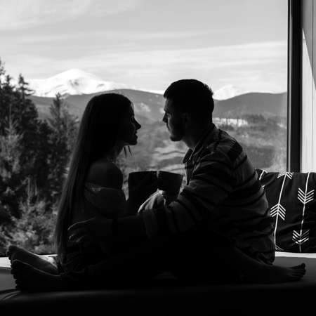 A young couple over a cup of coffee discussing everyday affairs, a guy and a girl sitting on the windowsill on a background of mountains and drinking coffee, relaxing in the Carpathians.の写真素材