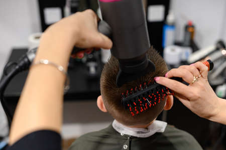 The hairdresser dries and styles the hair with the help of a hair dryer and a comb for a schoolboy boy, visits to the hairdresser during a coronavirus pandemic, black style hairdresser.の写真素材