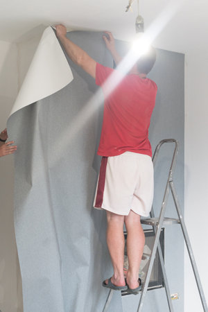 A man glues wallpaper on a wall in a house, a craftsman uses a ladder to glue wallpaper on top.