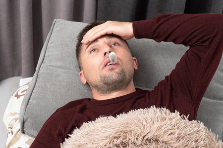 Man measures temperature and holds hand to head because of headache, sick man lies in bed.