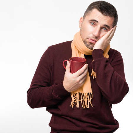 A handsome man touches his mouth with his hand with a pained expression because of a toothache or tooth disease, Dentist concept, a man wrapped in a scarf holds a mug in his hands.の写真素材